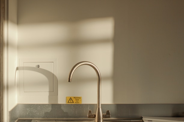 A minimalist kitchen scene featuring a sleek metallic faucet above a sink. Soft shadow patterns of a window are cast on the wall, creating a tranquil ambiance. A small yellow warning sign indicating 'very hot water' is visible near the faucet.