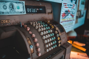 An antique cash register with numerous round, colorful keys is placed prominently. A vintage one-dollar bill is inserted above the display. The surroundings include posters and papers with colorful text and images.