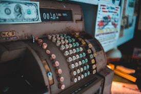 An antique cash register with numerous round, colorful keys is placed prominently. A vintage one-dollar bill is inserted above the display. The surroundings include posters and papers with colorful text and images.