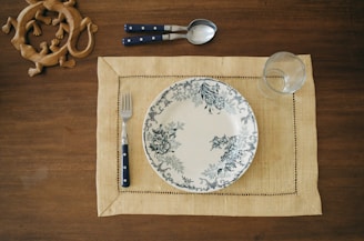 A neatly set table features a decorative plate with floral patterns on a woven placemat. Next to the plate, there is a fork and spoon with dark handles, and a clear glass is placed to the side. In the upper left corner, a wooden carving of intertwined lizards adds an artistic touch.