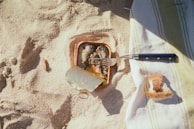 A sandy beach surface with a tin of sardines partially opened, showing fish inside. A fork rests on top of the tin, and nearby, on a striped beach towel, lies a piece of torn bread.