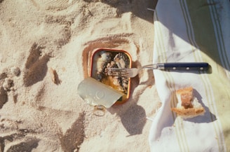 A sandy beach surface with a tin of sardines partially opened, showing fish inside. A fork rests on top of the tin, and nearby, on a striped beach towel, lies a piece of torn bread.