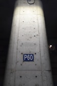 A concrete column with visible metal reinforcements and a rectangular blue sign reading 'P60'. The sign is centered on the column, which shows signs of weathering and dirt. The lighting highlights the center of the column, creating a contrast with the darker surroundings.