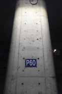 A concrete column with visible metal reinforcements and a rectangular blue sign reading 'P60'. The sign is centered on the column, which shows signs of weathering and dirt. The lighting highlights the center of the column, creating a contrast with the darker surroundings.