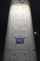 A concrete column with visible metal reinforcements and a rectangular blue sign reading 'P60'. The sign is centered on the column, which shows signs of weathering and dirt. The lighting highlights the center of the column, creating a contrast with the darker surroundings.