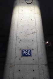 A concrete column with visible metal reinforcements and a rectangular blue sign reading 'P60'. The sign is centered on the column, which shows signs of weathering and dirt. The lighting highlights the center of the column, creating a contrast with the darker surroundings.