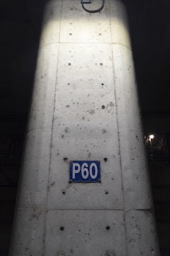 A concrete column with visible metal reinforcements and a rectangular blue sign reading 'P60'. The sign is centered on the column, which shows signs of weathering and dirt. The lighting highlights the center of the column, creating a contrast with the darker surroundings.