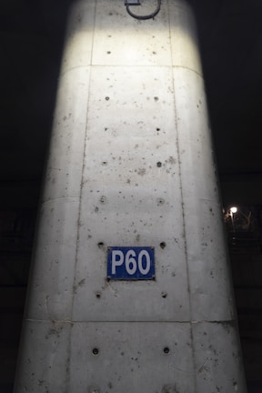 A concrete column with visible metal reinforcements and a rectangular blue sign reading 'P60'. The sign is centered on the column, which shows signs of weathering and dirt. The lighting highlights the center of the column, creating a contrast with the darker surroundings.