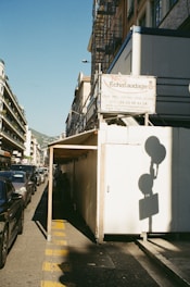 A street scene featuring a construction site with scaffolding on the side of a building. There is a covered walkway made of unfinished wood and white panels. The street is lined with parked cars and a sign in French is visible, advertising a scaffolding company. The sun casts long shadows and the sky is clear.