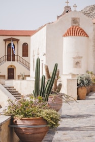 A peaceful courtyard with a beautiful combination of historical architecture and nature. A small chapel made of light stone with a red-tiled dome and crosses rests in the background. In front of the chapel, large potted cacti and other plants add greenery to the stone courtyard. A building with arched windows and a Greek flag hanging by the entrance complements the serene atmosphere.