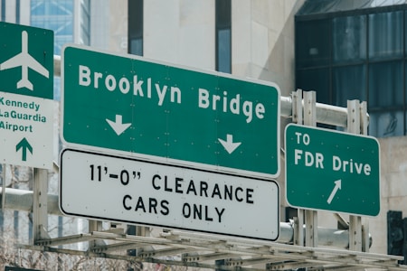 A group of green and white road signs guide drivers towards the Brooklyn Bridge, FDR Drive, and Kennedy/LaGuardia Airport. There is a height clearance restriction for cars indicated on one of the signs.