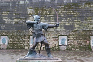 A bronze statue of an archer in a traditional outfit, drawing a bow, placed in front of a moss-covered stone wall. The figure is standing on a raised platform, wearing a tunic with a belt and a hat with a feather. Behind the statue, the wall has several stone plaques with faint reliefs engraved on them.