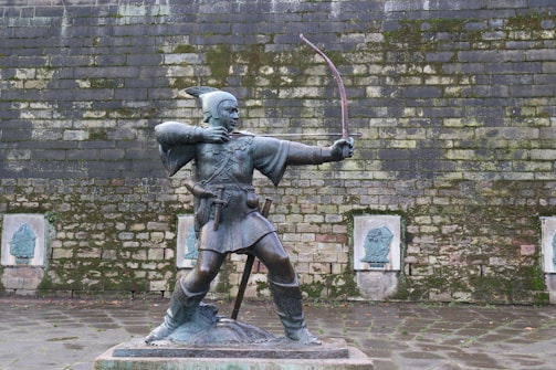 A bronze statue of an archer in a traditional outfit, drawing a bow, placed in front of a moss-covered stone wall. The figure is standing on a raised platform, wearing a tunic with a belt and a hat with a feather. Behind the statue, the wall has several stone plaques with faint reliefs engraved on them.