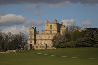 A grand historic mansion stands majestically surrounded by lush greenery. The architecture features intricate detailing with towers and large windows, typical of traditional European styles. A vast expanse of manicured lawn stretches in the foreground, while clouds hover in the blue sky.