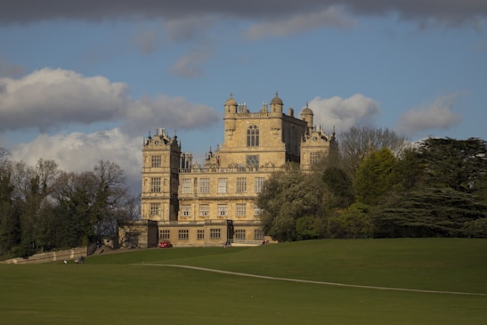 A grand historic mansion stands majestically surrounded by lush greenery. The architecture features intricate detailing with towers and large windows, typical of traditional European styles. A vast expanse of manicured lawn stretches in the foreground, while clouds hover in the blue sky.