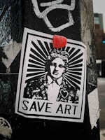 A monochromatic street art poster is pasted on a weathered surface, featuring a person in a suit with a stylized sunburst design emanating from their head. Above the head is a small red object, possibly a candy or sticker. The poster includes the text 'SAVE ART' at the bottom and a heart symbol on the individual’s jacket.