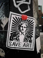 A monochromatic street art poster is pasted on a weathered surface, featuring a person in a suit with a stylized sunburst design emanating from their head. Above the head is a small red object, possibly a candy or sticker. The poster includes the text 'SAVE ART' at the bottom and a heart symbol on the individual’s jacket.
