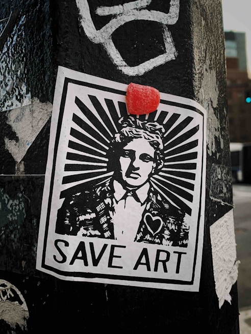 A monochromatic street art poster is pasted on a weathered surface, featuring a person in a suit with a stylized sunburst design emanating from their head. Above the head is a small red object, possibly a candy or sticker. The poster includes the text 'SAVE ART' at the bottom and a heart symbol on the individual’s jacket.