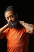 A bearded man with curly hair is holding his hands behind his neck. He is wearing an orange t-shirt with the quote 'I Sell Digital Dope' printed on it, along with a tag '@shotbysham.' The background is solid black, creating a stark contrast with the subject's attire. The man's expression appears relaxed and casual.