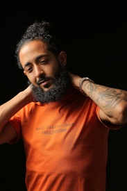 A bearded man with curly hair is holding his hands behind his neck. He is wearing an orange t-shirt with the quote 'I Sell Digital Dope' printed on it, along with a tag '@shotbysham.' The background is solid black, creating a stark contrast with the subject's attire. The man's expression appears relaxed and casual.