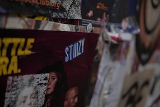A collection of layered posters and advertisements, including partially visible text with the word 'STIIIZY' prominently displayed in blue. The images on the posters feature various people and abstract designs, with some sections obscured or overlapping. The overall scene is busy and textured, conveying a sense of urban culture.