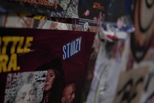 A collection of layered posters and advertisements, including partially visible text with the word 'STIIIZY' prominently displayed in blue. The images on the posters feature various people and abstract designs, with some sections obscured or overlapping. The overall scene is busy and textured, conveying a sense of urban culture.