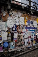 A busy urban wall is covered with a colorful collage of posters, graffiti, and street art. Messages such as 'Be Kind' are prominently displayed, along with various artistic drawings and event advertisements. The overall appearance is layered and textured, conveying a sense of vibrant urban culture.