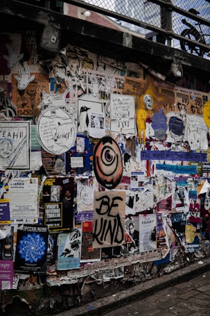 A busy urban wall is covered with a colorful collage of posters, graffiti, and street art. Messages such as 'Be Kind' are prominently displayed, along with various artistic drawings and event advertisements. The overall appearance is layered and textured, conveying a sense of vibrant urban culture.