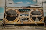 A large mural painted on the side of a building depicts a vintage-style boombox with the words 'Everlasting Bass' prominently displayed. The artwork features two large speaker designs and various dials and buttons. A person stands in front of the mural, adding a sense of scale.