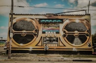 A large mural painted on the side of a building depicts a vintage-style boombox with the words 'Everlasting Bass' prominently displayed. The artwork features two large speaker designs and various dials and buttons. A person stands in front of the mural, adding a sense of scale.