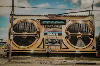 A large mural painted on the side of a building depicts a vintage-style boombox with the words 'Everlasting Bass' prominently displayed. The artwork features two large speaker designs and various dials and buttons. A person stands in front of the mural, adding a sense of scale.