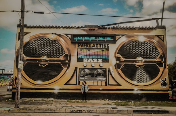 A large mural painted on the side of a building depicts a vintage-style boombox with the words 'Everlasting Bass' prominently displayed. The artwork features two large speaker designs and various dials and buttons. A person stands in front of the mural, adding a sense of scale.