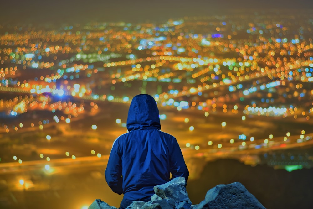 person wearing blue hooded jacket standing on rocky hill viewing city during night time