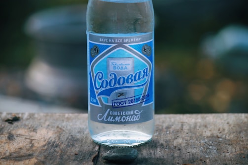 A clear glass bottle with a blue and white label stands on a wooden surface. The label contains Cyrillic text and decorative designs, giving it a vintage appearance. The background is blurred, focusing attention on the bottle.