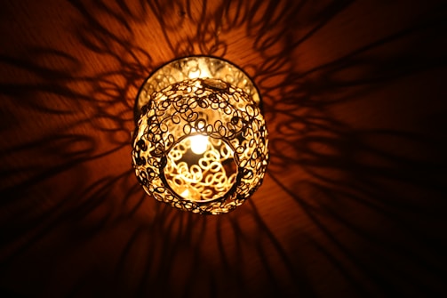 A decorative light fixture is mounted on the ceiling, featuring an intricate metal design that casts complex shadows onto the surrounding surface. The light radiates a warm, golden glow, enhancing the artistic patterns created by the metal loops.