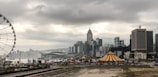 An urban landscape featuring a city skyline with a high concentration of skyscrapers. In the foreground, a large, striped circus tent with yellow and red colors is visible. To the left, a Ferris wheel stands tall, adding an element of amusement. The sky is overcast with thick clouds, creating a dramatic atmosphere.