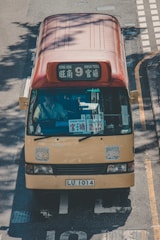 A yellow minibus with a red roof traveling on a road. The front display shows destinations including 'Mong Kok' and 'Kwun Tong'. Several stickers are visible on the windshield and body. The vehicle's license plate reads 'LU 1014'. The scene is illuminated by natural sunlight, casting shadows on the road.