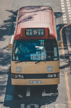 A yellow minibus with a red roof traveling on a road. The front display shows destinations including 'Mong Kok' and 'Kwun Tong'. Several stickers are visible on the windshield and body. The vehicle's license plate reads 'LU 1014'. The scene is illuminated by natural sunlight, casting shadows on the road.