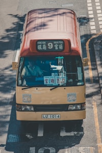 A yellow minibus with a red roof traveling on a road. The front display shows destinations including 'Mong Kok' and 'Kwun Tong'. Several stickers are visible on the windshield and body. The vehicle's license plate reads 'LU 1014'. The scene is illuminated by natural sunlight, casting shadows on the road.