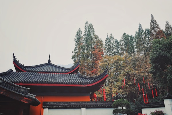 A traditional East Asian architectural structure with an ornate, curved roof is surrounded by dense greenery. Red lanterns are hung in the trees, contrasting with the muted colors of the foliage and building. The scene conveys a sense of cultural heritage and tranquility.