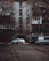A residential apartment building with a weathered facade, featuring multiple windows and balconies. Several vehicles, including a silver car and a gray van, are parked in front of the building on an empty street. Leafless trees frame the scene, contributing to an overall urban and slightly neglected appearance.