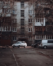 A residential apartment building with a weathered facade, featuring multiple windows and balconies. Several vehicles, including a silver car and a gray van, are parked in front of the building on an empty street. Leafless trees frame the scene, contributing to an overall urban and slightly neglected appearance.