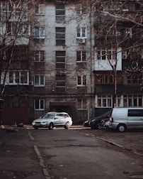 A residential apartment building with a weathered facade, featuring multiple windows and balconies. Several vehicles, including a silver car and a gray van, are parked in front of the building on an empty street. Leafless trees frame the scene, contributing to an overall urban and slightly neglected appearance.