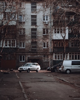 A residential apartment building with a weathered facade, featuring multiple windows and balconies. Several vehicles, including a silver car and a gray van, are parked in front of the building on an empty street. Leafless trees frame the scene, contributing to an overall urban and slightly neglected appearance.