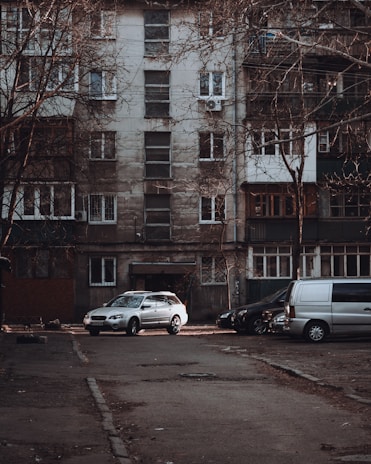 A residential apartment building with a weathered facade, featuring multiple windows and balconies. Several vehicles, including a silver car and a gray van, are parked in front of the building on an empty street. Leafless trees frame the scene, contributing to an overall urban and slightly neglected appearance.