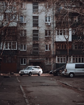 A residential apartment building with a weathered facade, featuring multiple windows and balconies. Several vehicles, including a silver car and a gray van, are parked in front of the building on an empty street. Leafless trees frame the scene, contributing to an overall urban and slightly neglected appearance.