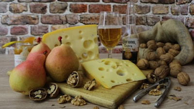 A rustic still life arrangement featuring pears, slices of Swiss cheese, walnuts, and a glass of beer on a wooden cutting board. There is also a jar of honey, a bottle of beer, and a burlap sack partially filled with walnuts. The background consists of a brick wall, adding a cozy and warm atmosphere to the setting.