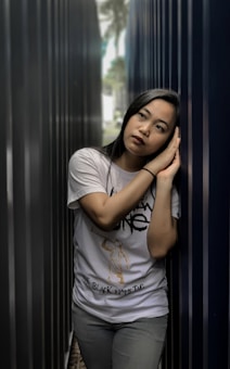 A person is leaning against a metal wall in a narrow alley. The individual appears to be in deep thought, with their head resting on their hands. The background is out of focus, highlighting the person's contemplative expression. The lighting is soft, creating a calm and introspective atmosphere.