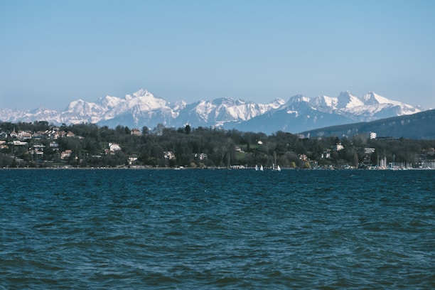 A serene lake with gentle waves in the foreground, surrounded by lush greenery and residential houses on the shoreline. In the background, majestic snow-capped mountains stretch across the horizon under a clear blue sky.