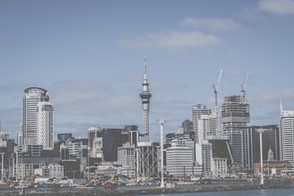 A modern city skyline featuring a mix of high-rise buildings, including a prominent tower with a slender structure. Construction cranes are visible, indicating ongoing development. The sky is partly cloudy, and the waterfront is lined with various structures.